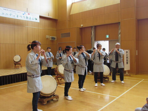 太鼓が置かれた体育館の中で、法被を着た人々が笛を吹いている写真