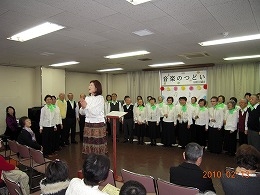 小さなホールの中、「音楽のつどい」の横断幕を背にして、大勢の人々が合唱をしている写真