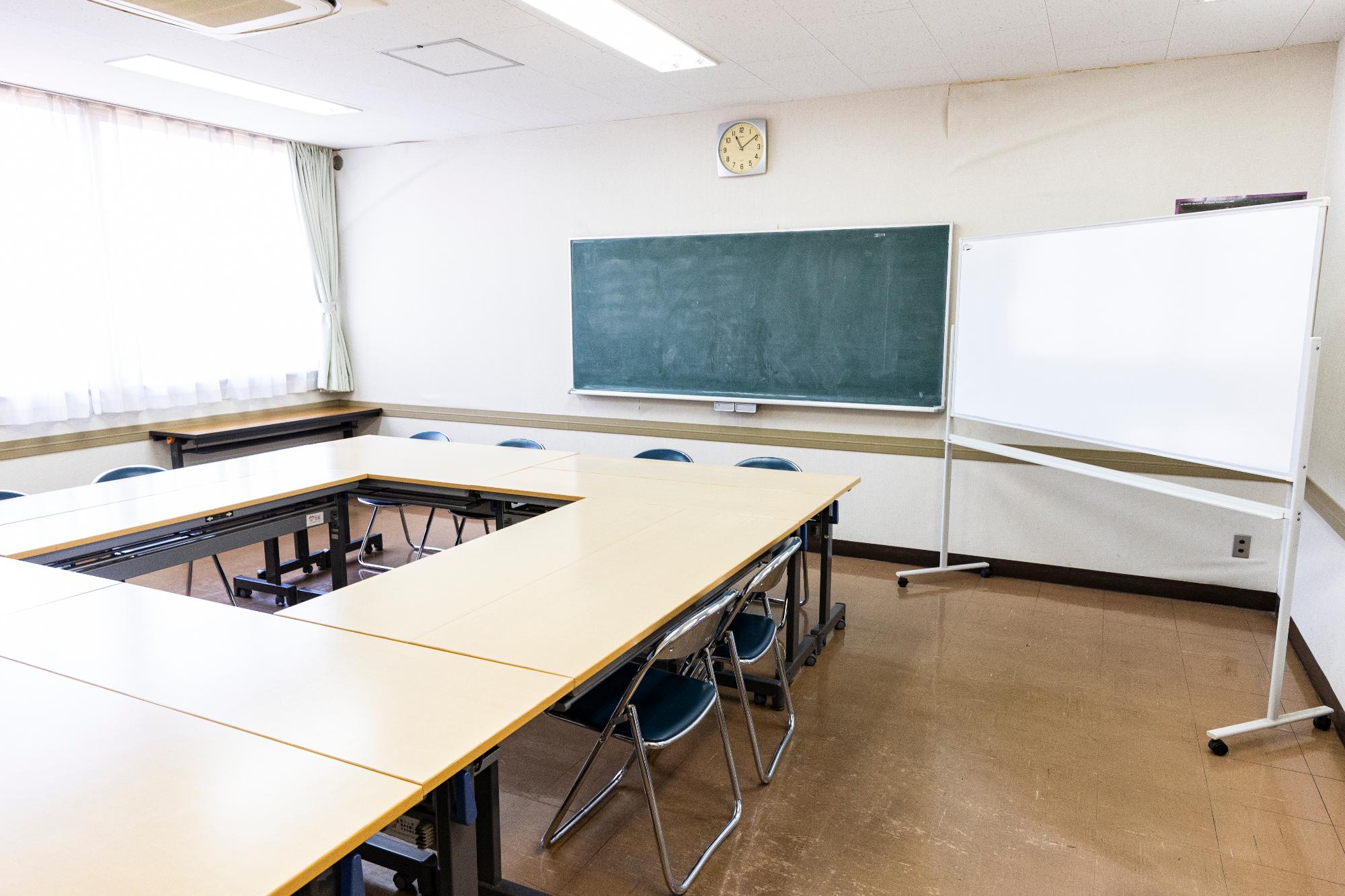 窓から光が入る部屋の中、正面に黒板とホワイトボードが置かれ、机が中央を空けて置かれている写真