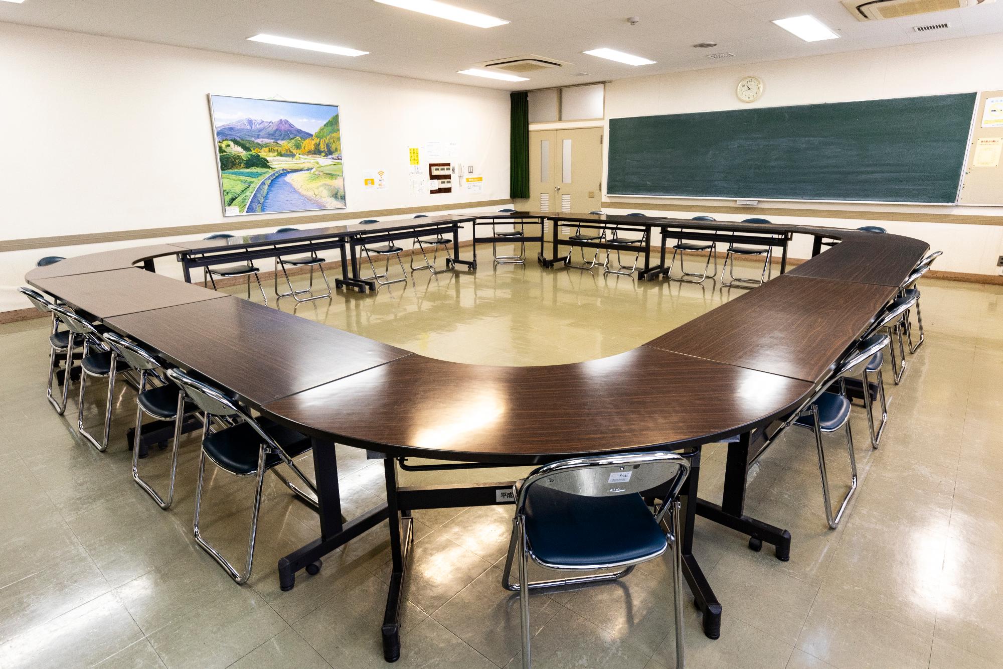 黒板と絵画が置かれた部屋の中、机と椅子が中央を空けて設置されている写真