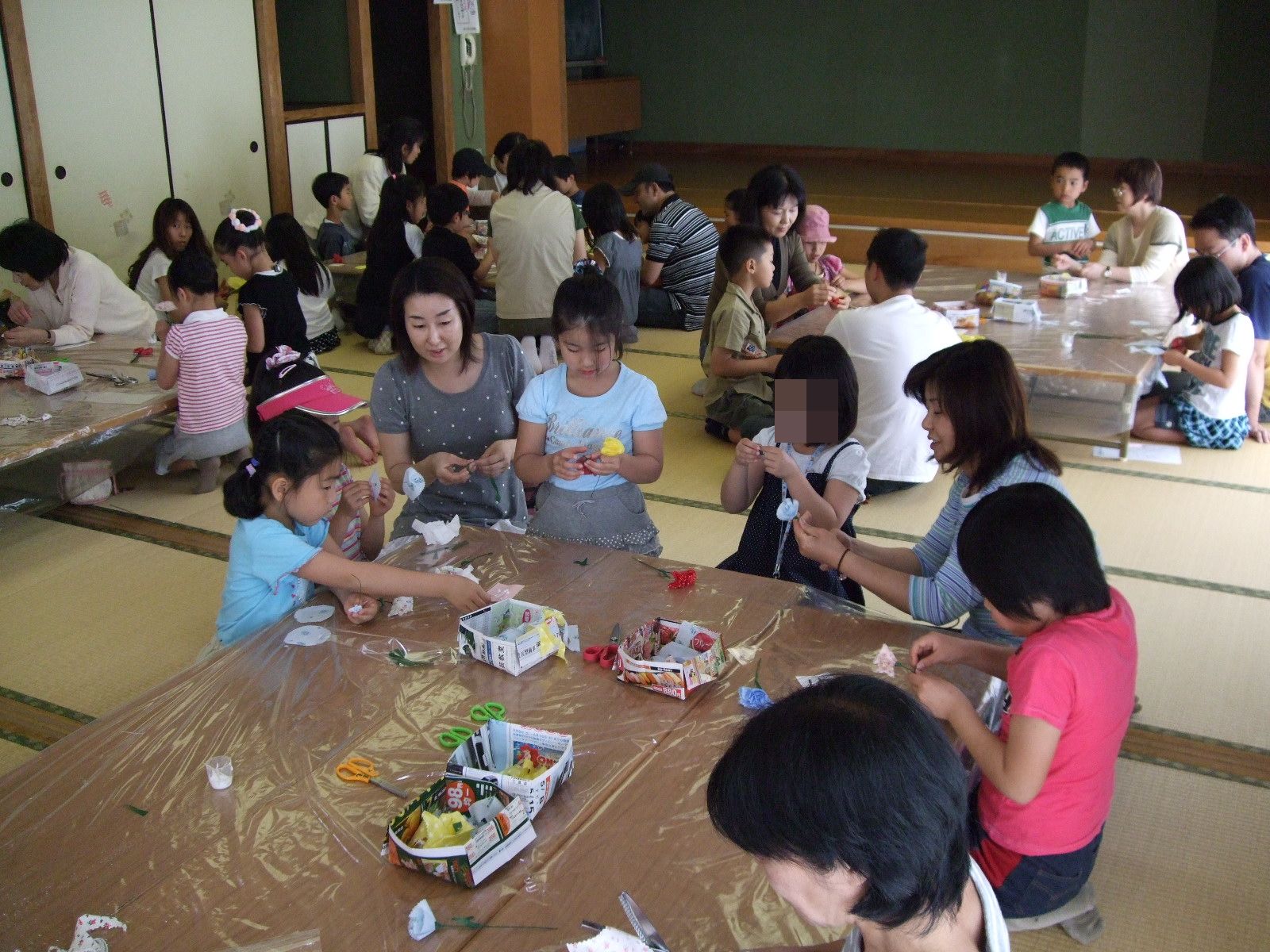 和室に座り、机を並べてテープのカーネーションを作る大勢の親子の写真
