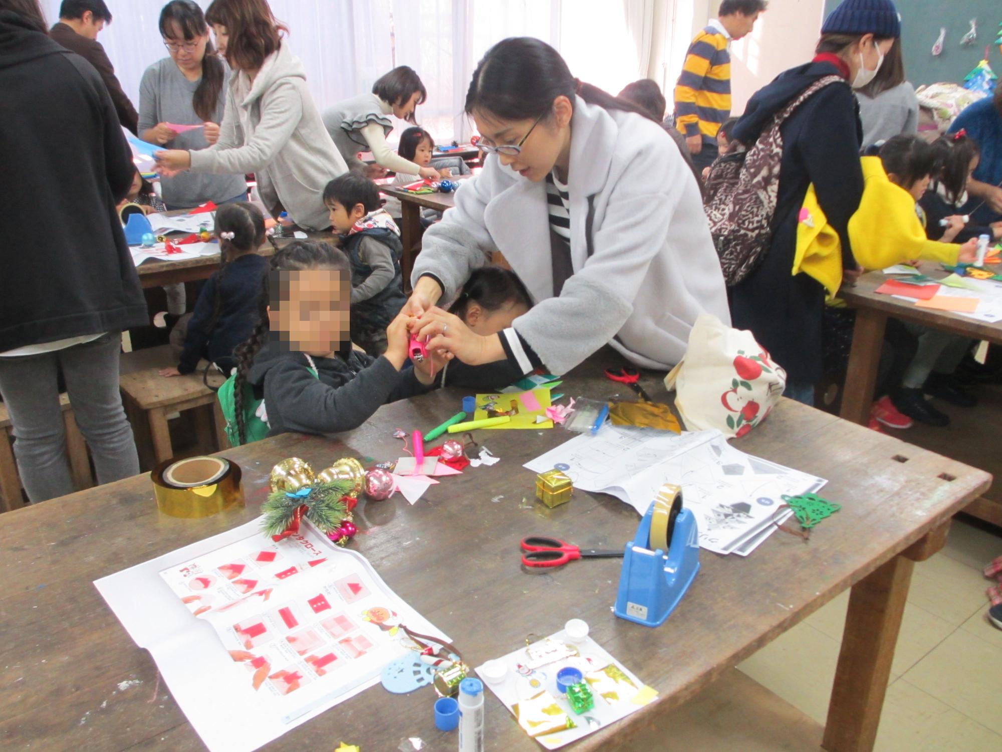 沢山の机の中の1つに注目した、折り紙を楽しむ子供二人と保護者の写真