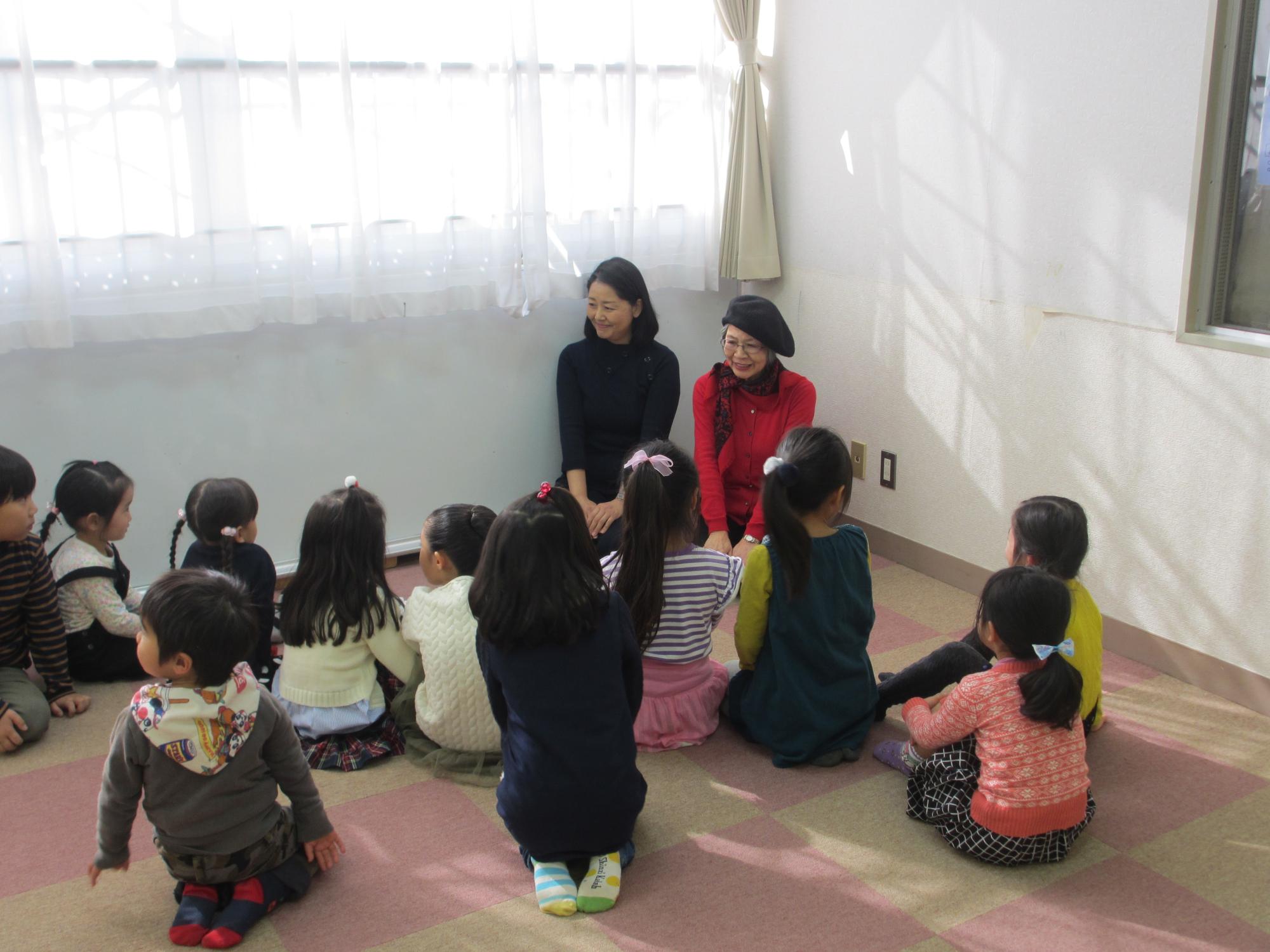 部屋の隅で二人の大人と大勢の子供たちが、地面に座りながら話している写真