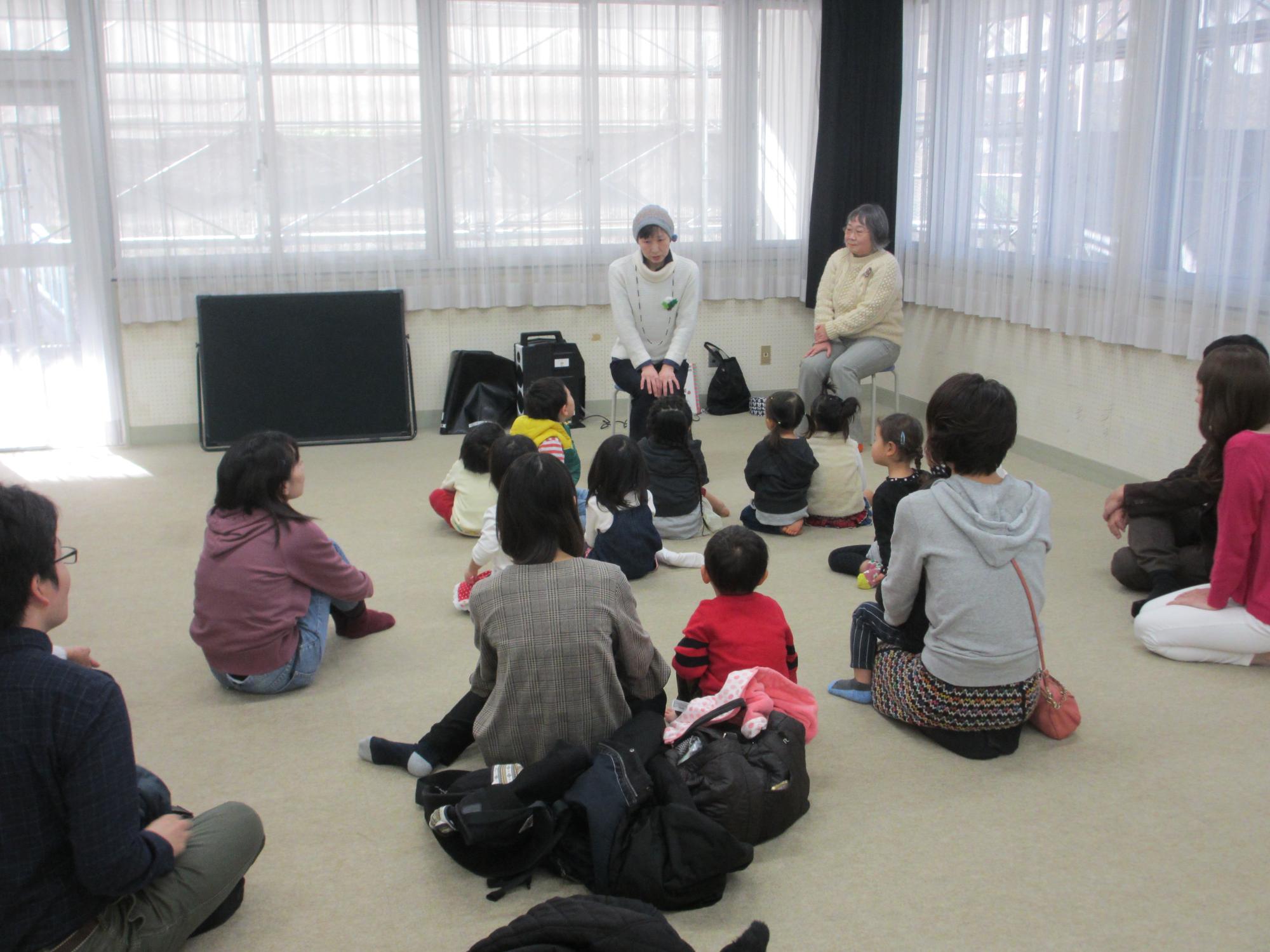 2人の大人と大勢の子供たちが座りながら話しているところを、後ろから写した写真