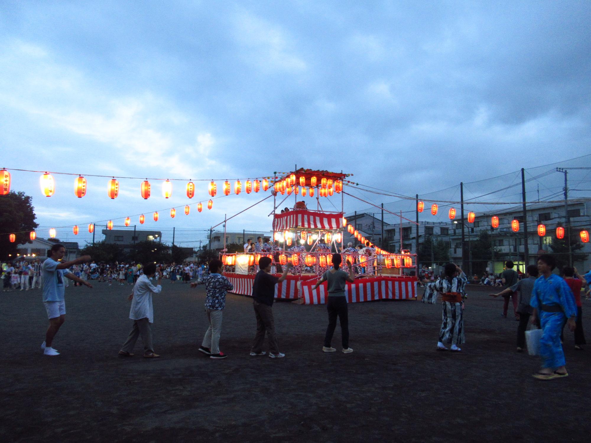 暗くなってきた空の下で、赤色の提灯で飾られたやぐらと周囲で盆踊りを楽しむ人々の写真