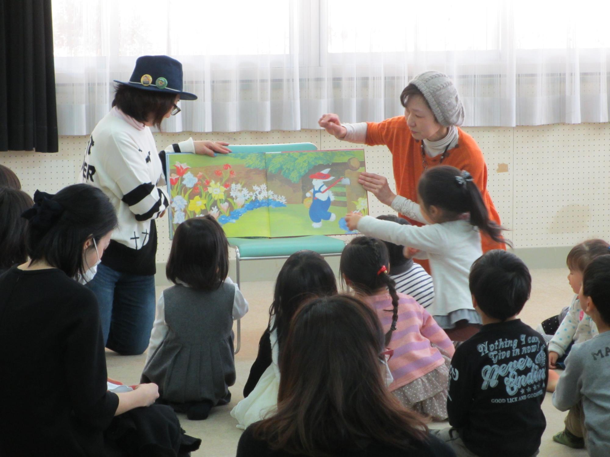 絵本の読み聞かせを行う二人の女性と、近くで集まって見ている子ども達の写真