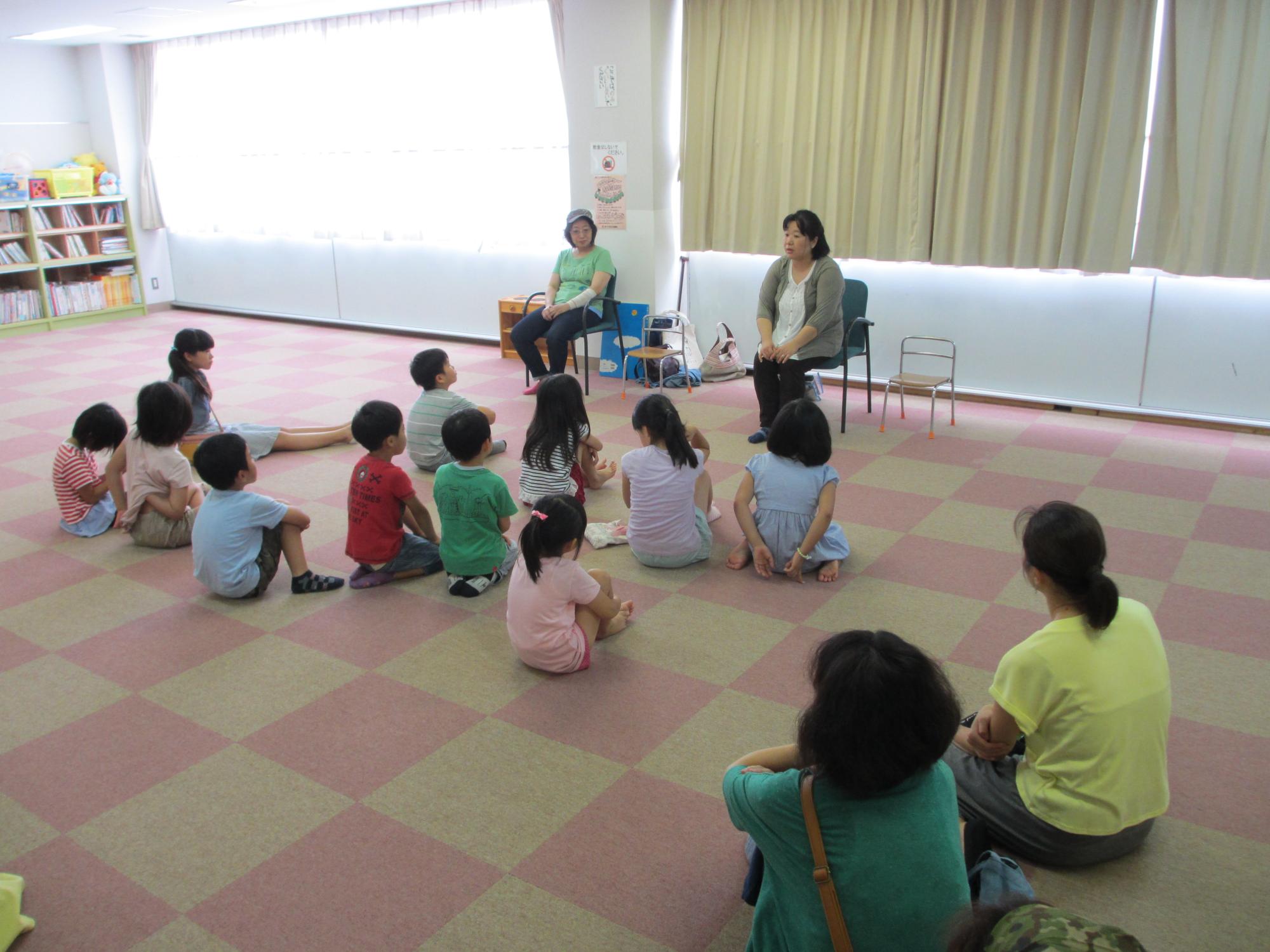 椅子に座って話している二人の女性とそれを見ている大勢の子供たち、見守る二人の大人の写真