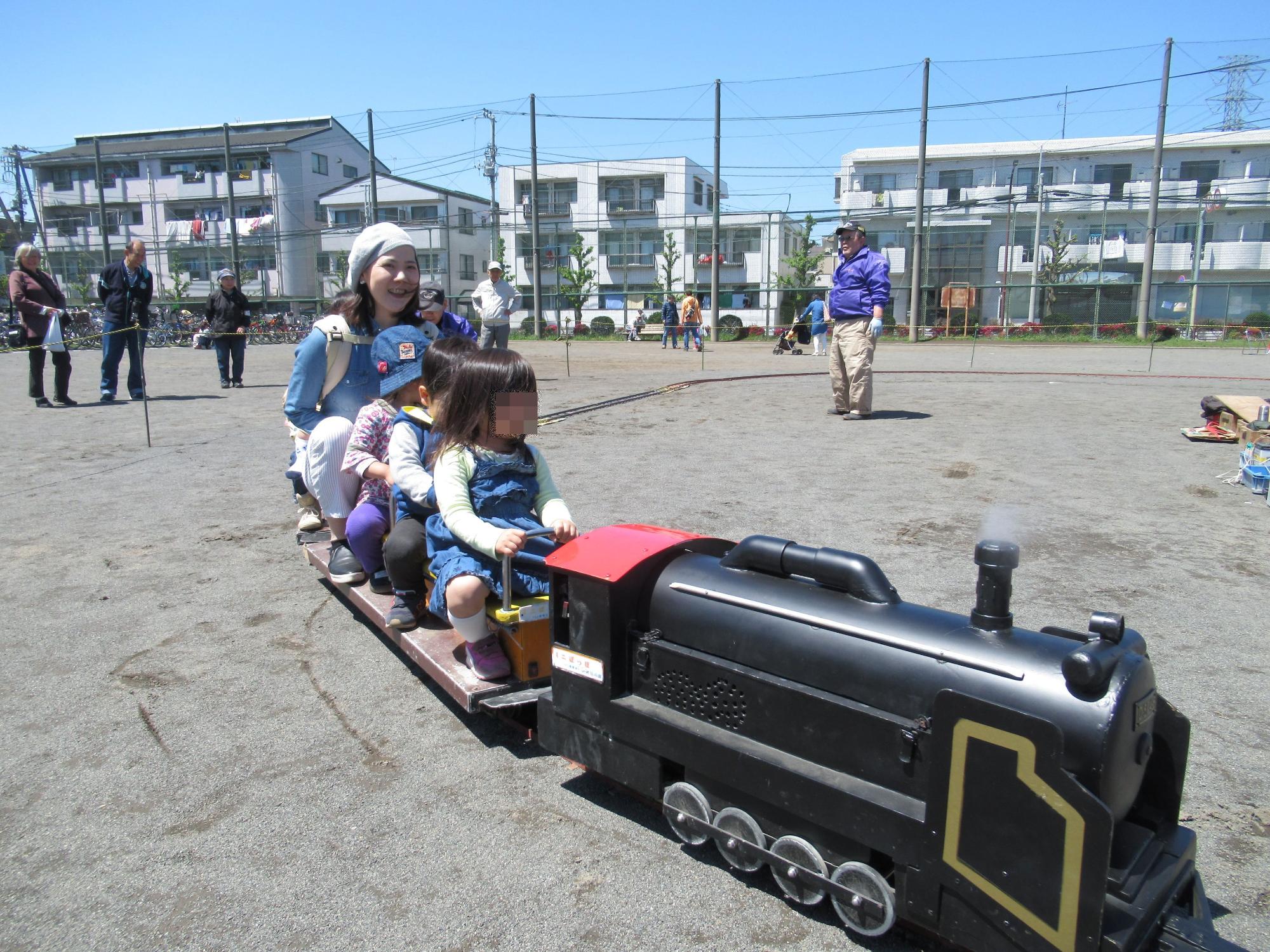 校庭で列車型の遊具に乗って遊ぶ二人の子供と保護者の写真