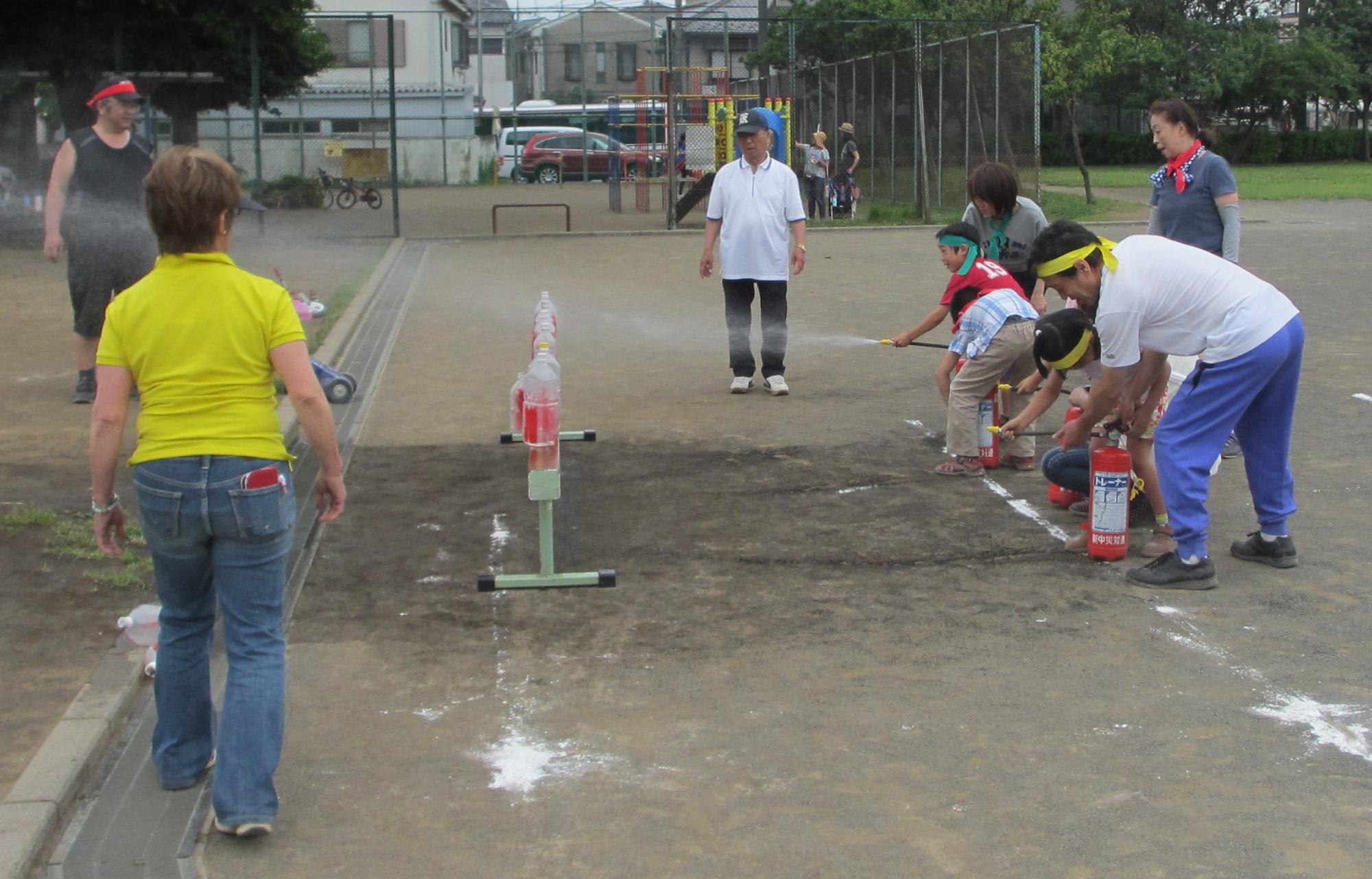 校庭で消火器を使い、ペットボトルに拭きかけている、二人の子どもと協力する二人の大人の写真