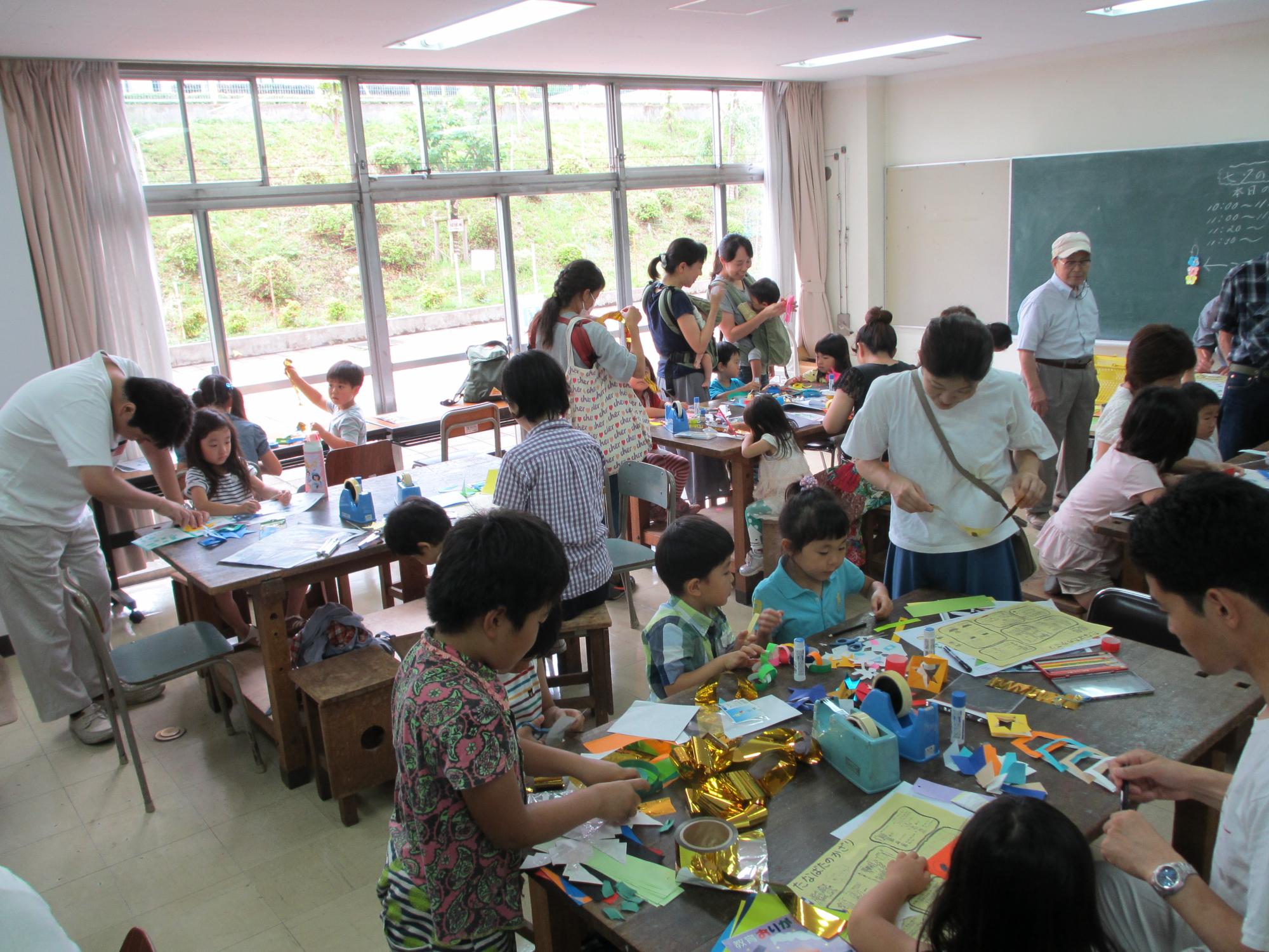 教室の中で四つの机にそれぞれ向かい、紙で飾りを作る子供たちと見守る複数の大人の写真