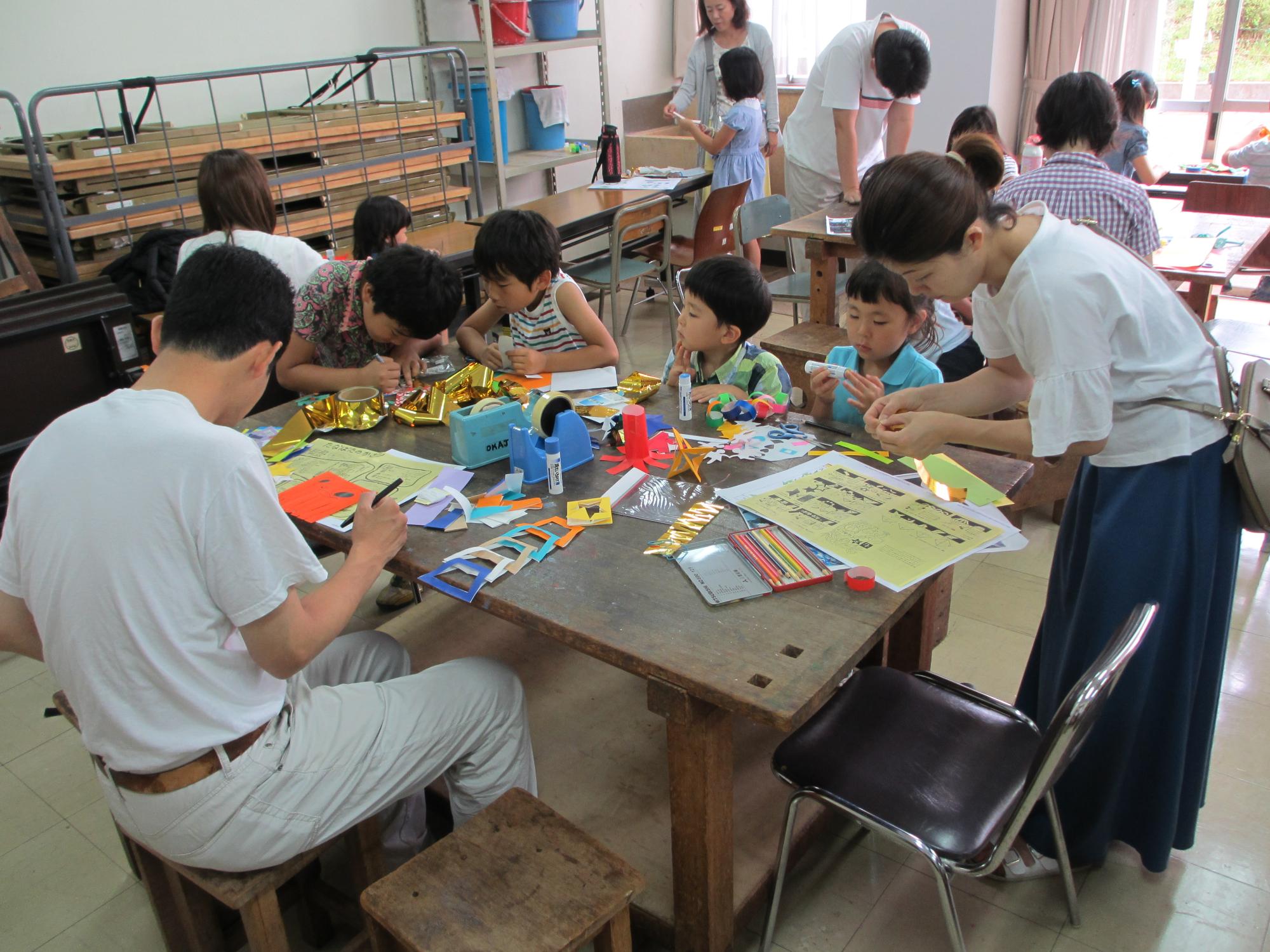 複数ある机の1つに注目した、紙で飾りを作る四人の子供と二人の大人の写真