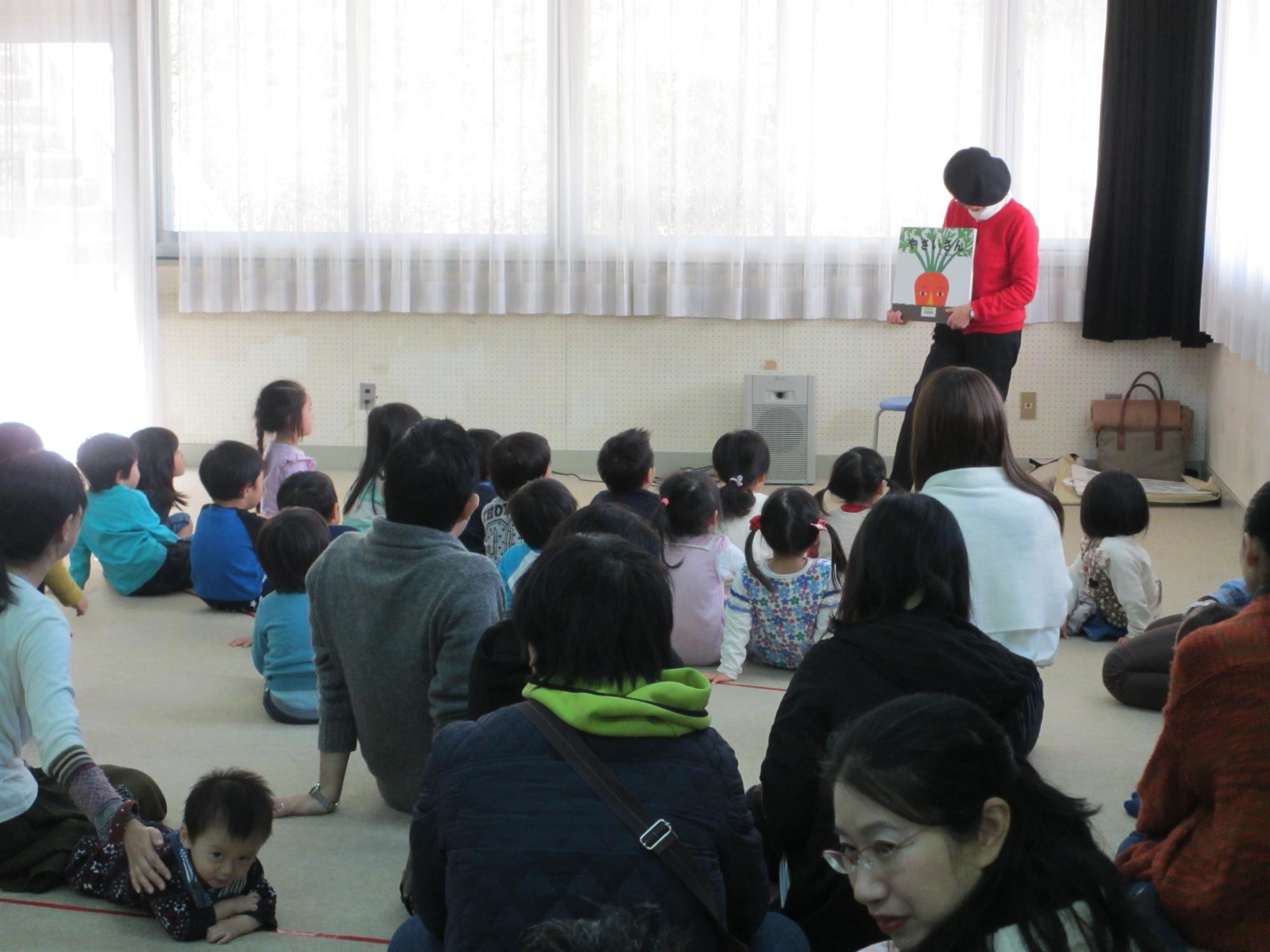 絵本の読み聞かせをする大人と、それを見ている大勢の子供たちの写真