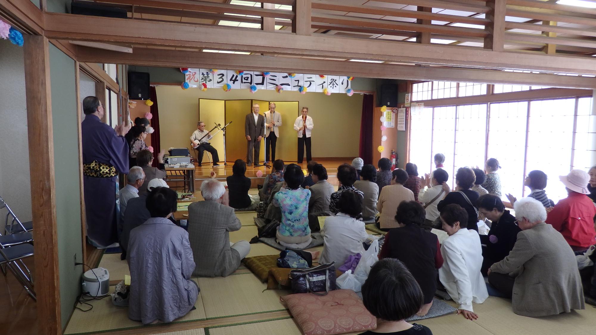 「第34回コミュニティ祭り」の幕の下で、演奏をする四人の男性と座って観覧する多くの人々の写真