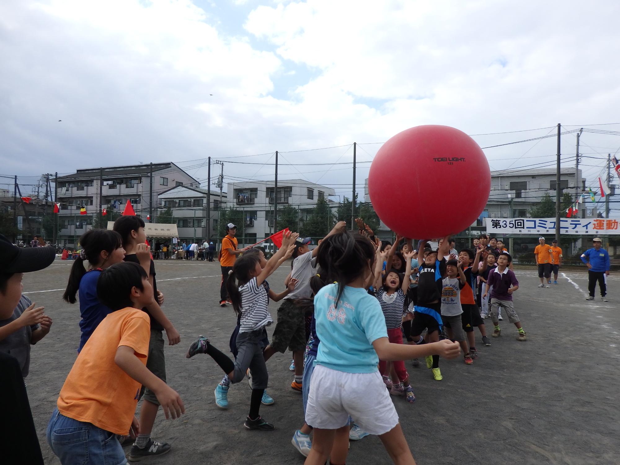 赤色の大きな球を頭の上で転がしていく子どもたちの写真