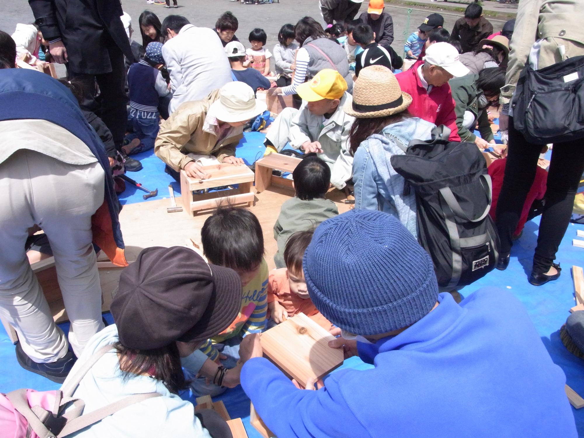 校庭の下で大勢の人々が集まる中で、数人の人々が木箱を研磨している写真