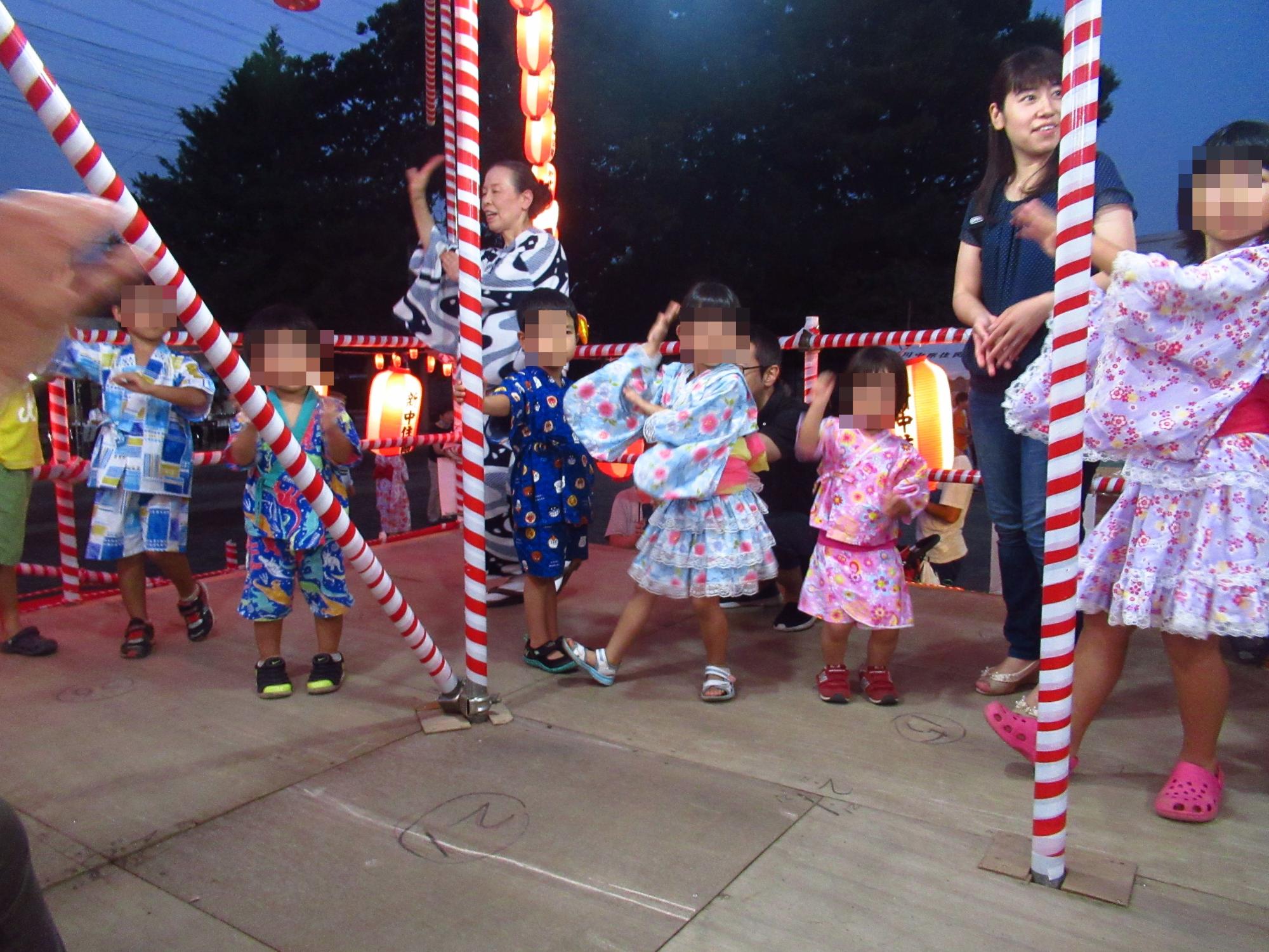 夜にやぐらの上で盆踊りを楽しむ、六人の子供と二人の大人の写真