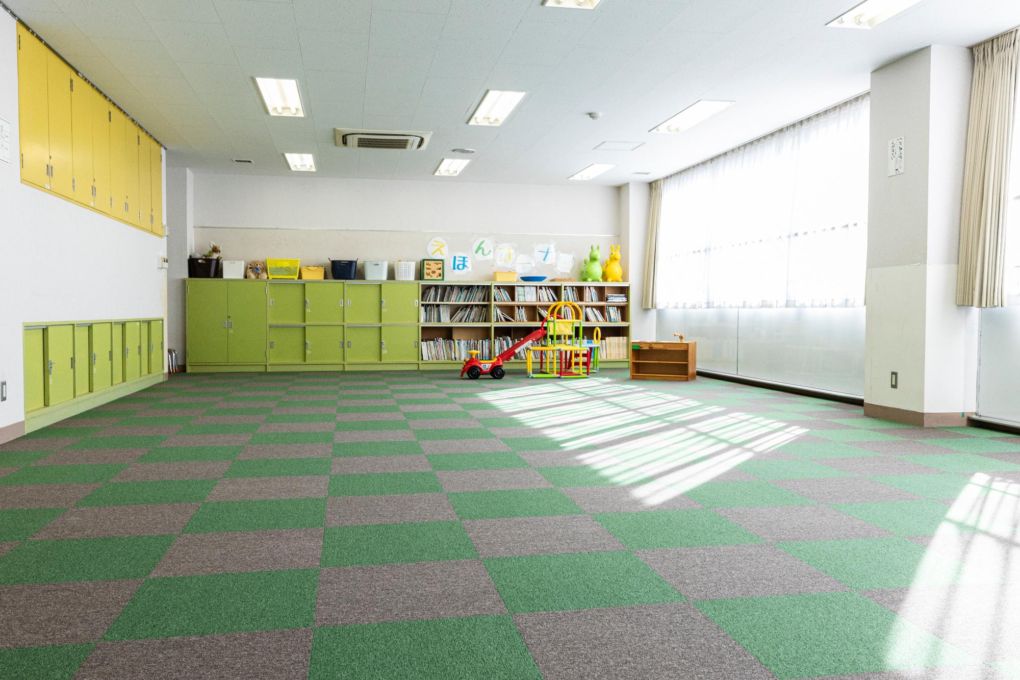 床に緑と灰色のカーペットが貼られ、周囲に緑色と黄色のロッカーと絵本やおもちゃが多数用意されている幼児が使える部屋の写真